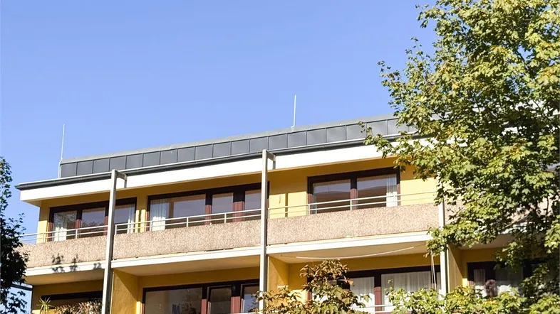 Fassade eines Mehrfamilienhauses mit Balkonen und grüner Umgebung unter blauem Himmel.