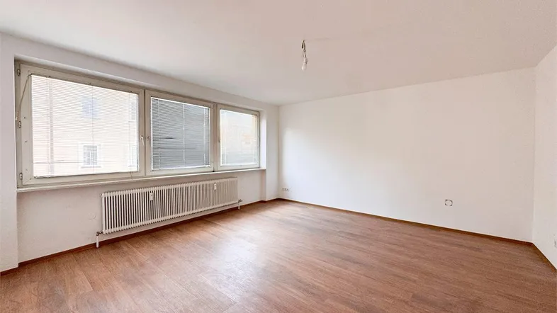 Helles Zimmer mit Holzfußboden, weißen Wänden und großen Fenstern mit Jalousien.