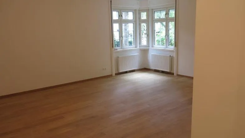 Helles, leeres Zimmer mit Parkettboden und einem Erkerfenster, das viel Tageslicht hereinlässt.