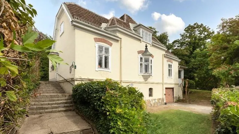 Klassisches Gebäude mit heller Fassade, Ziegeldach und einer Steintreppe, umgeben von üppigem Grün und Bäumen.