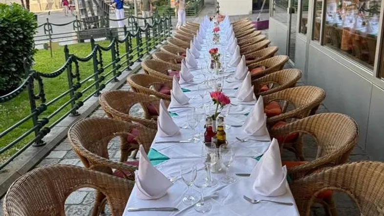 Lange, festlich gedeckte Tafel auf einer Außenterrasse mit Rattanmöbeln und Blick auf die Straße.