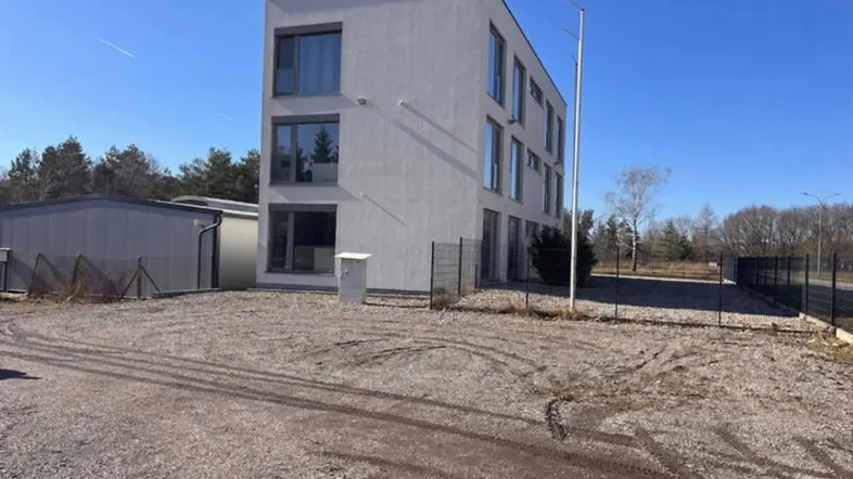 Modernes, weißes Gebäude mit großen Fenstern und einem Schotterparkplatz unter blauem Himmel.