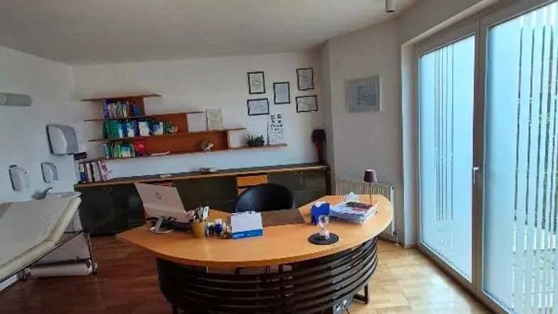 Helles Büro oder Behandlungszimmer mit großem Fenster, Holzfußboden und modernem Schreibtisch.