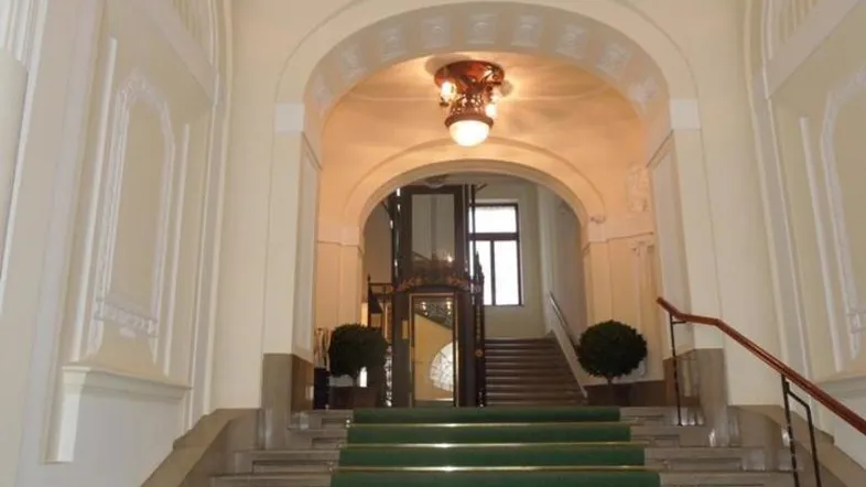 Elegantes Treppenhaus mit grünem Teppich und einem historischen Aufzug hinter einem Bogen.