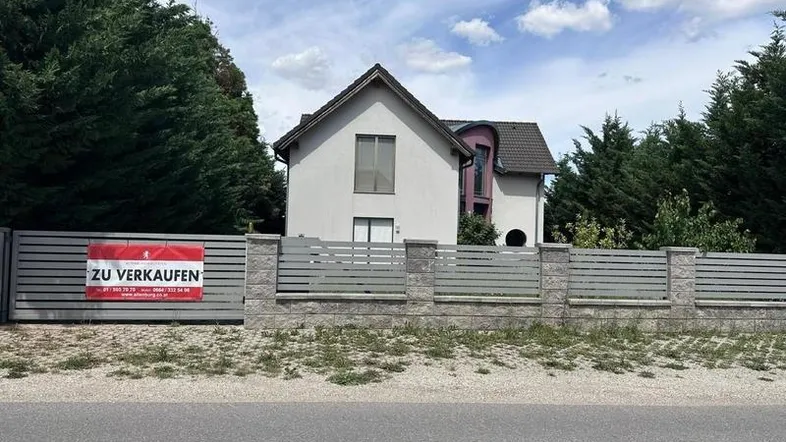 Modernes Einfamilienhaus mit Zaun und "Zu Verkaufen"-Schild im Vordergrund.