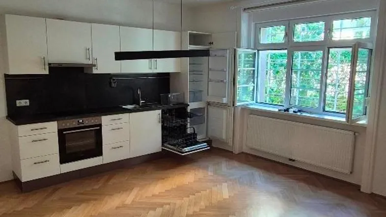 Moderne Einbauküche mit weißen Schränken, dunkler Arbeitsplatte und Fischgrätparkett in einem hellen Raum.