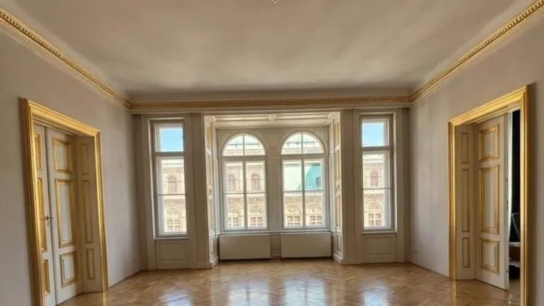 Geräumiges Wohnzimmer mit Fischgrätparkett, hohen Decken und vergoldeten Stuckelementen.