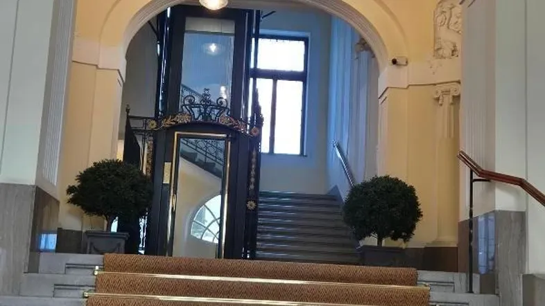 Elegantes Treppenhaus mit Teppichläufer, Stuckdecke und einem gläsernen Aufzug in einem Altbau.