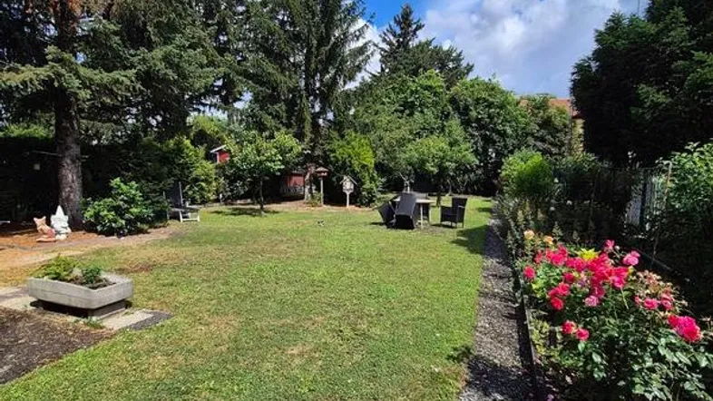 Gepflegter Garten mit Rasenfläche, hohen Bäumen und blühenden Rosenbeeten unter blauem Himmel.