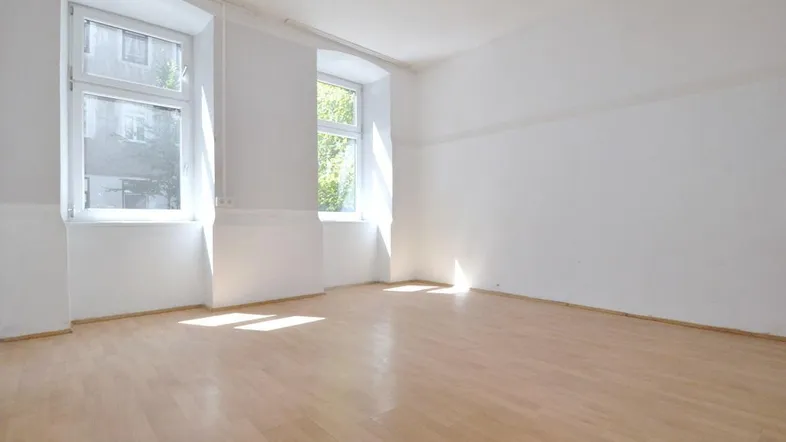 Heller, leerer Raum mit Holzboden und zwei Fenstern, die viel Tageslicht hereinlassen.