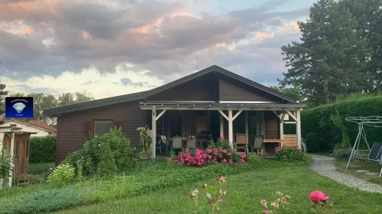 Gemütliches Holzhaus mit überdachter Terrasse und blühendem Garten unter einem bewölkten Himmel.