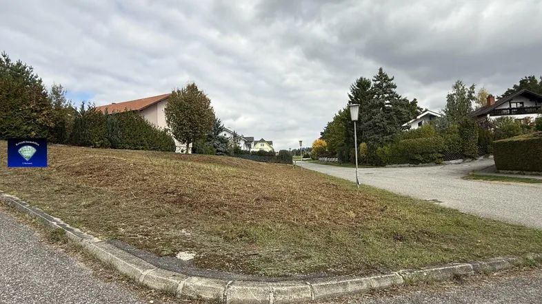 Leeres Grundstück an einer Straßenecke mit umliegenden Wohnhäusern unter bewölktem Himmel.