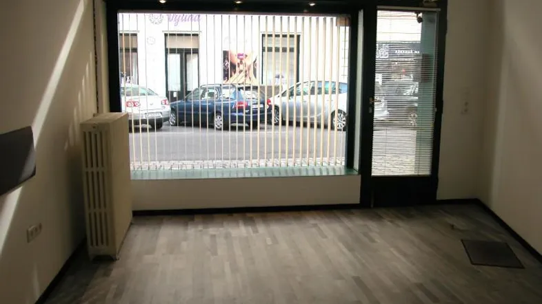Leerer Raum mit großen Fenstern zur Straße, ausgestattet mit einem Heizkörper und Holzplankenboden.