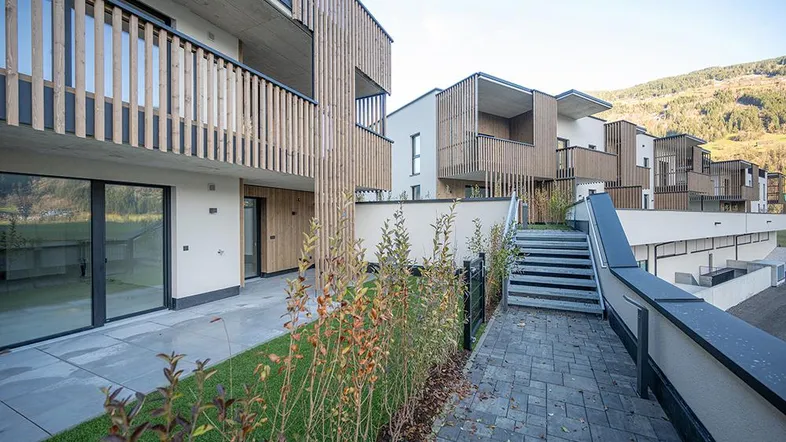 Moderne Wohnanlage mit Holzfassade, Balkonen und privaten Gärten im Erdgeschoss, umgeben von Berglandschaft.