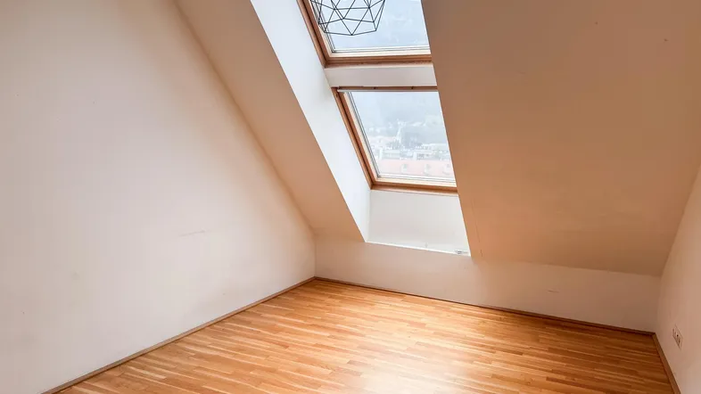 Helles Zimmer im Dachgeschoss mit Holzboden und zwei großen Dachfenstern, die viel Tageslicht hereinlassen.