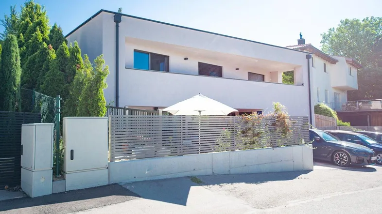 Moderne Hausfassade mit Terrasse, Sonnenschirm und Parkplätzen vor dem Gebäude.