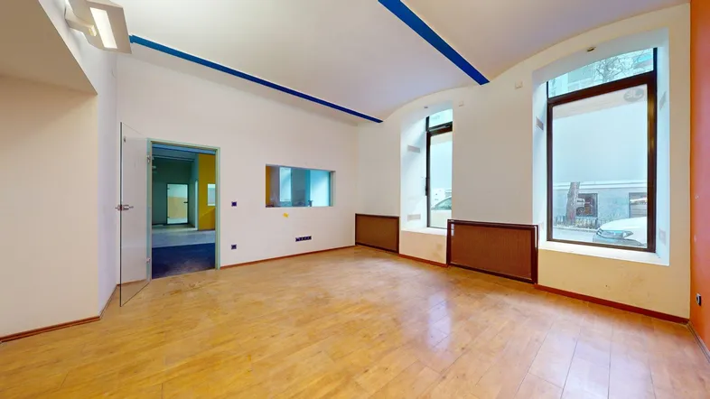 Geräumiger, leerer Raum mit Holzboden, großen Fenstern und blauen Akzentstreifen an der Decke.