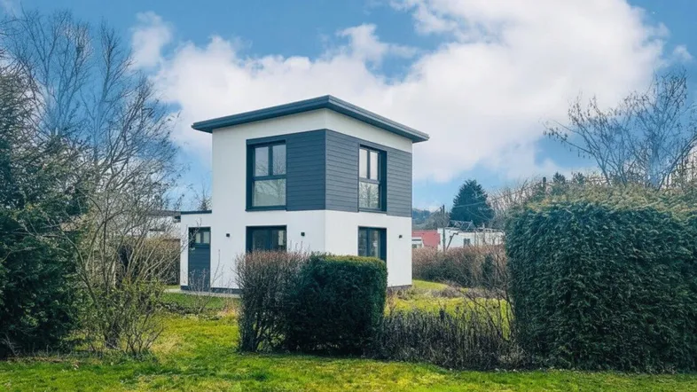 Modernes Einfamilienhaus mit weißer Fassade und dunklen Akzenten, umgeben von Grünflächen unter blauem Himmel.