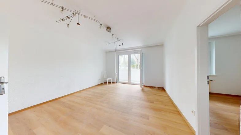 Helles, leeres Wohnzimmer mit Parkettboden und Zugang zu einem Balkon.