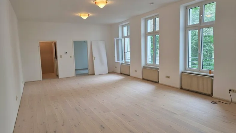 Heller, leerer Raum mit neu verlegtem Holzboden und mehreren hohen Fenstern, die viel Tageslicht hereinlassen.