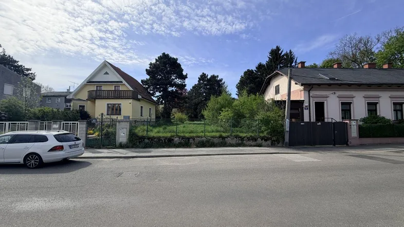 Blick von der Straße auf ein unbebautes Grundstück mit umliegenden Häusern und Bäumen.