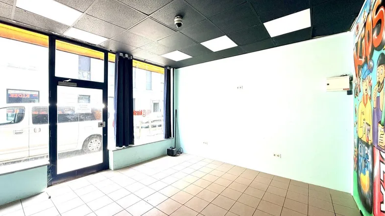 Leerer Ladenraum mit großen Schaufenstern, Fliesenboden und einer bunten Wandmalerei an der Seite.