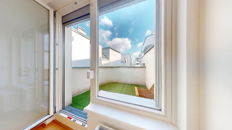 Blick aus einem hellen Raum auf eine kleine begrünte Terrasse mit Holzsessel.