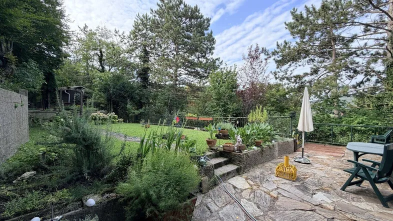 Große Steinterrasse mit Gartenmöbeln, Sonnenschirm und üppiger Bepflanzung, umgeben von Bäumen.