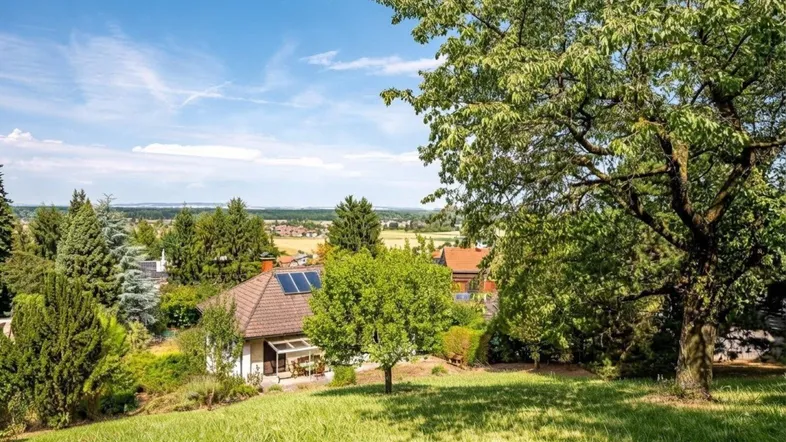 Weitläufiger Blick über die grüne Landschaft mit einem Haus und Bäumen unter blauem Himmel.
