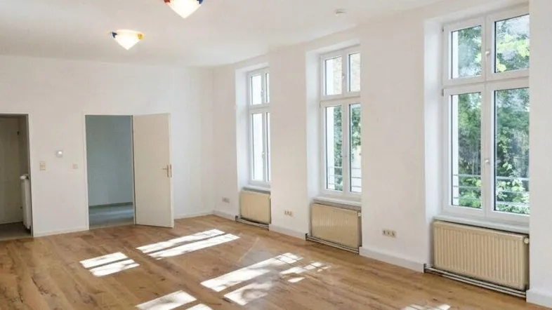 Heller, leerer Wohnraum mit Holzboden, hohen Fenstern und viel natürlichem Licht.