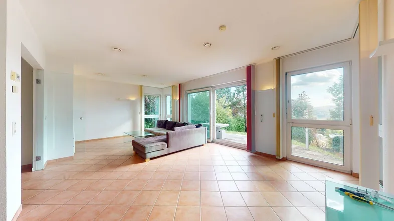 Helles Wohnzimmer mit Fliesenboden, großzügigen Fenstern und Zugang zu einer Terrasse mit Grünblick.