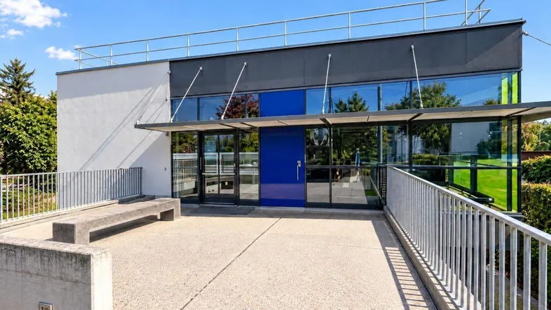Modernes Gebäude mit Glasfassade und blauem Eingangsbereich, umgeben von einer gepflasterten Terrasse.