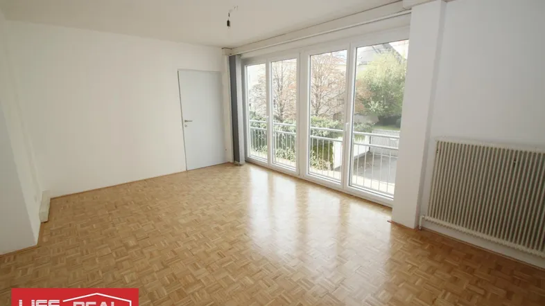 Heller Wohnraum mit Fischgrätparkett, großen Fenstern und Zugang zu einem Balkon mit Blick ins Grüne.