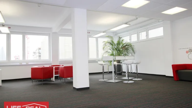 Großer, offener Bürobereich mit roten Sitzgelegenheiten, hohen Tischen und vielen Fenstern für natürliches Licht.