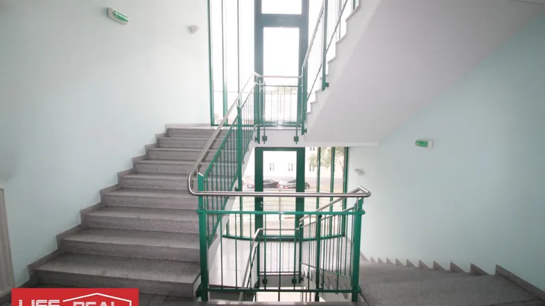 Helles Treppenhaus mit mehreren Etagen, grünen Geländern und großen Fenstern für viel Tageslicht.