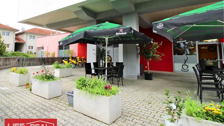 Außenbereich mit Tischen, Stühlen und Sonnenschirmen, umgeben von Blumenkübeln und einem roten Gebäude.