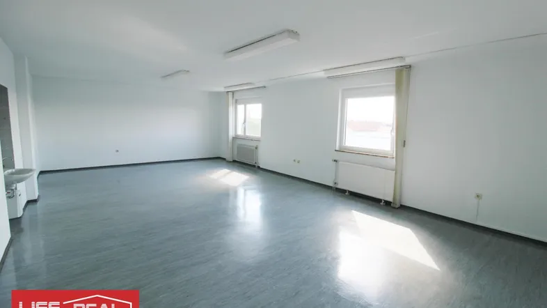Heller, leerer Büroraum mit großen Fenstern, Vinylboden und einem Waschbecken in der Ecke.