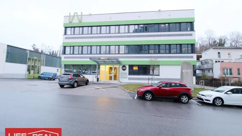 Modernes Bürogebäude mit grünen Akzenten und Parkplätzen davor, an einem bewölkten Tag.