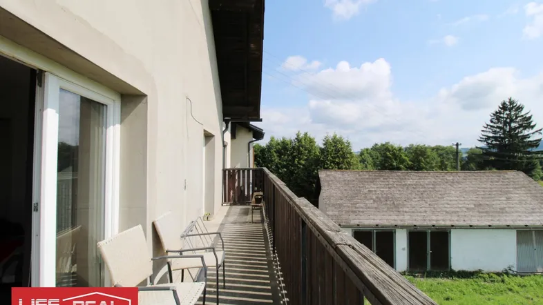 Geräumiger Balkon mit Holzboden und Geländer, bietet einen weiten Blick auf die umliegende Landschaft und ein Nebengebäude.