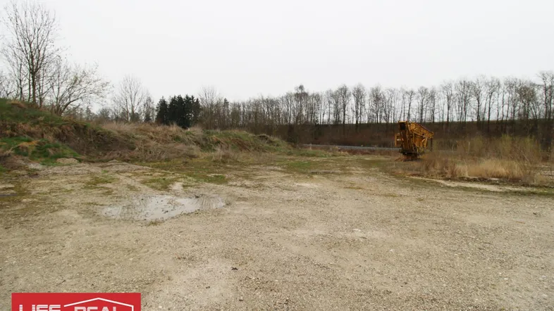 Weitläufiges, ungenutztes Grundstück mit Schotterfläche und Baumbestand im Hintergrund unter bewölktem Himmel.