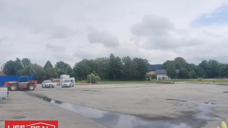 Großer, leerer Grundstücksbereich mit befestigter Fläche und Bäumen im Hintergrund unter bewölktem Himmel.