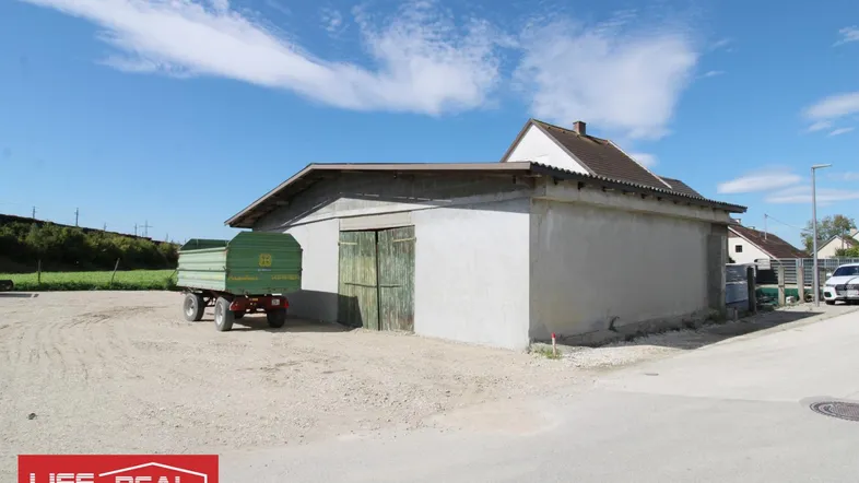 Ein landwirtschaftliches Gebäude mit einem Anhänger davor, umgeben von Schotter und einem blauen Himmel.