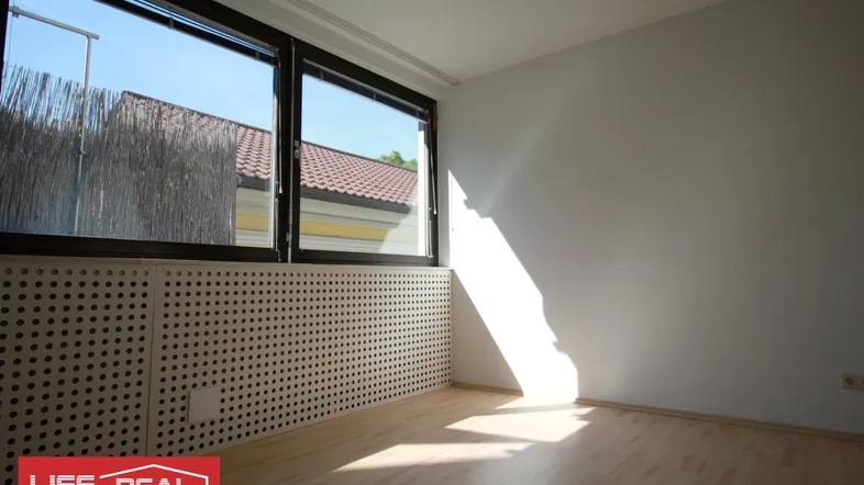 Heller Raum mit großen Fenstern, Holzboden und einem Heizkörper, der von Sonnenlicht durchflutet wird.
