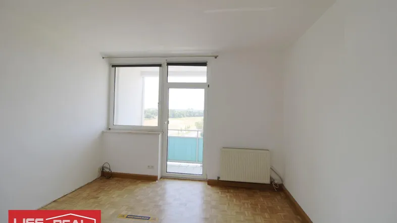 Leerer Raum mit Parkettboden, Heizkörper und Zugang zu einem kleinen Balkon mit Blick ins Grüne.