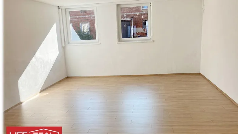 Leerer Raum mit hellem Laminatboden und weißen Wänden, durchflutet von natürlichem Sonnenlicht.