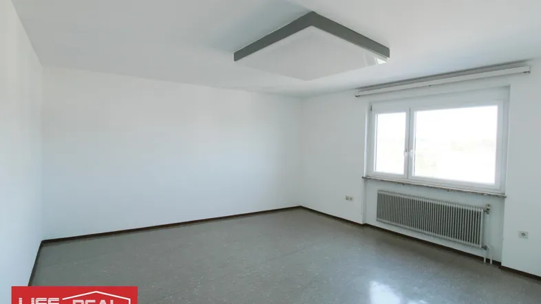 Heller, leerer Raum mit weißen Wänden, grauem Boden und einem Fenster mit Heizkörper darunter.