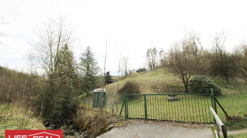 Grünes Metalltor führt zu einem Grundstück mit Bachlauf, grasbewachsenen Hügeln und Bäumen.