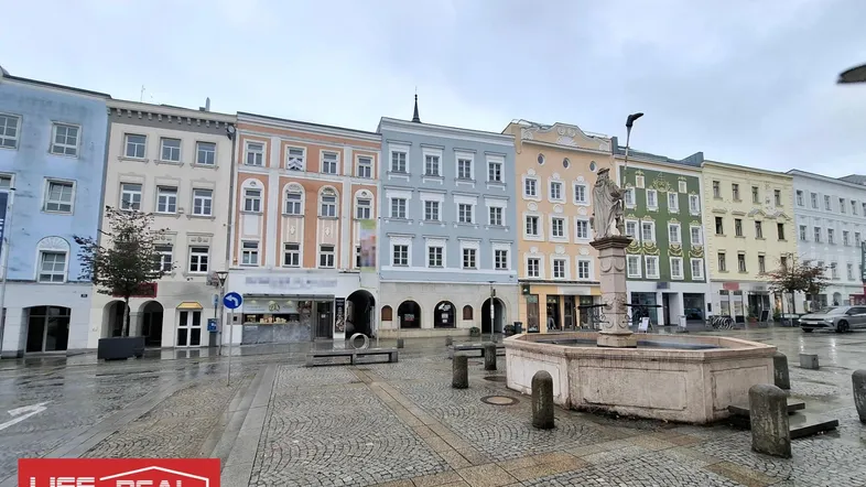 Historischer Stadtplatz mit bunten Gebäuden und einem Brunnen unter bewölktem Himmel.