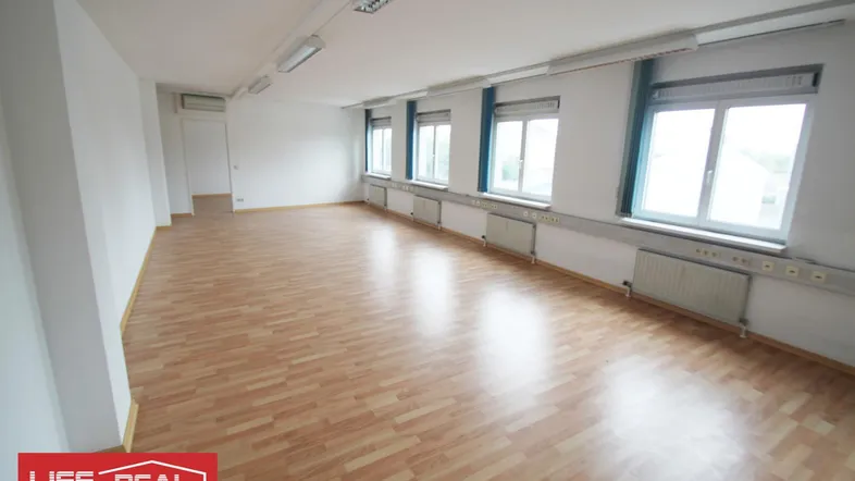 Geräumiger, leerer Büroraum mit hellem Laminatboden und mehreren Fenstern, die viel Tageslicht hereinlassen.