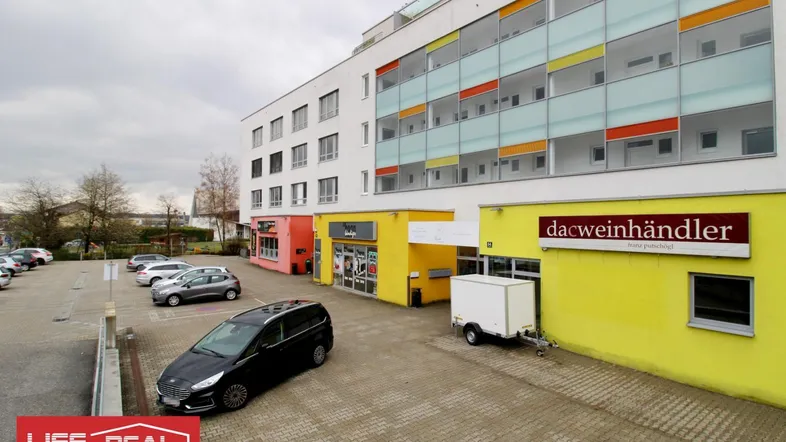 Modernes Geschäftsgebäude mit farbigen Akzenten und Außenparkplätzen unter bewölktem Himmel.